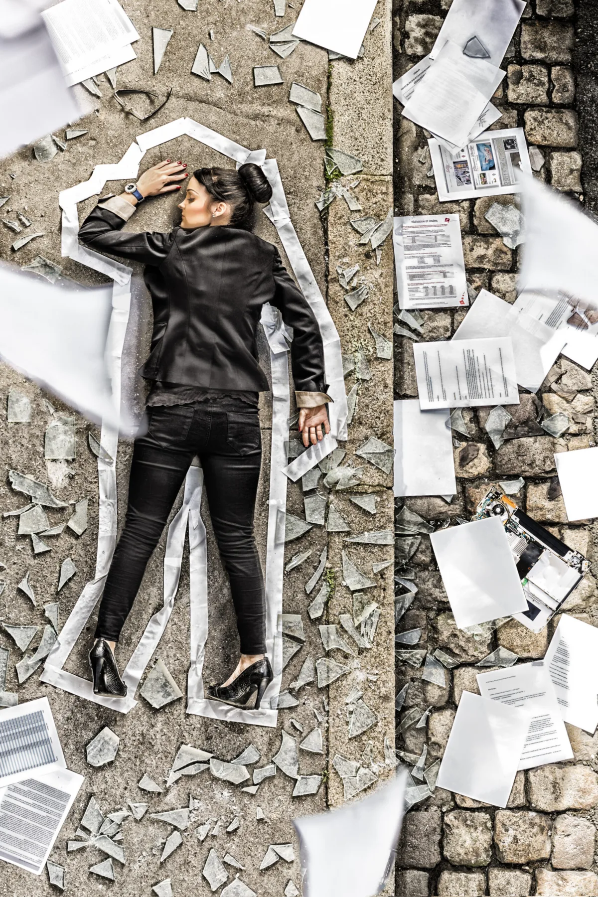 A woman lies on cobblestones inside a chalk outline, papers scattered around her