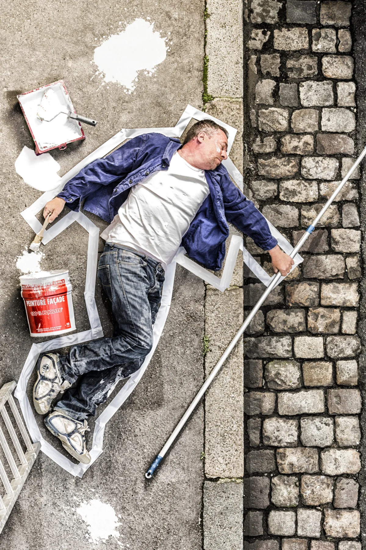 A painter lies on cobblestones inside a chalk outline, paint pot and roller beside him