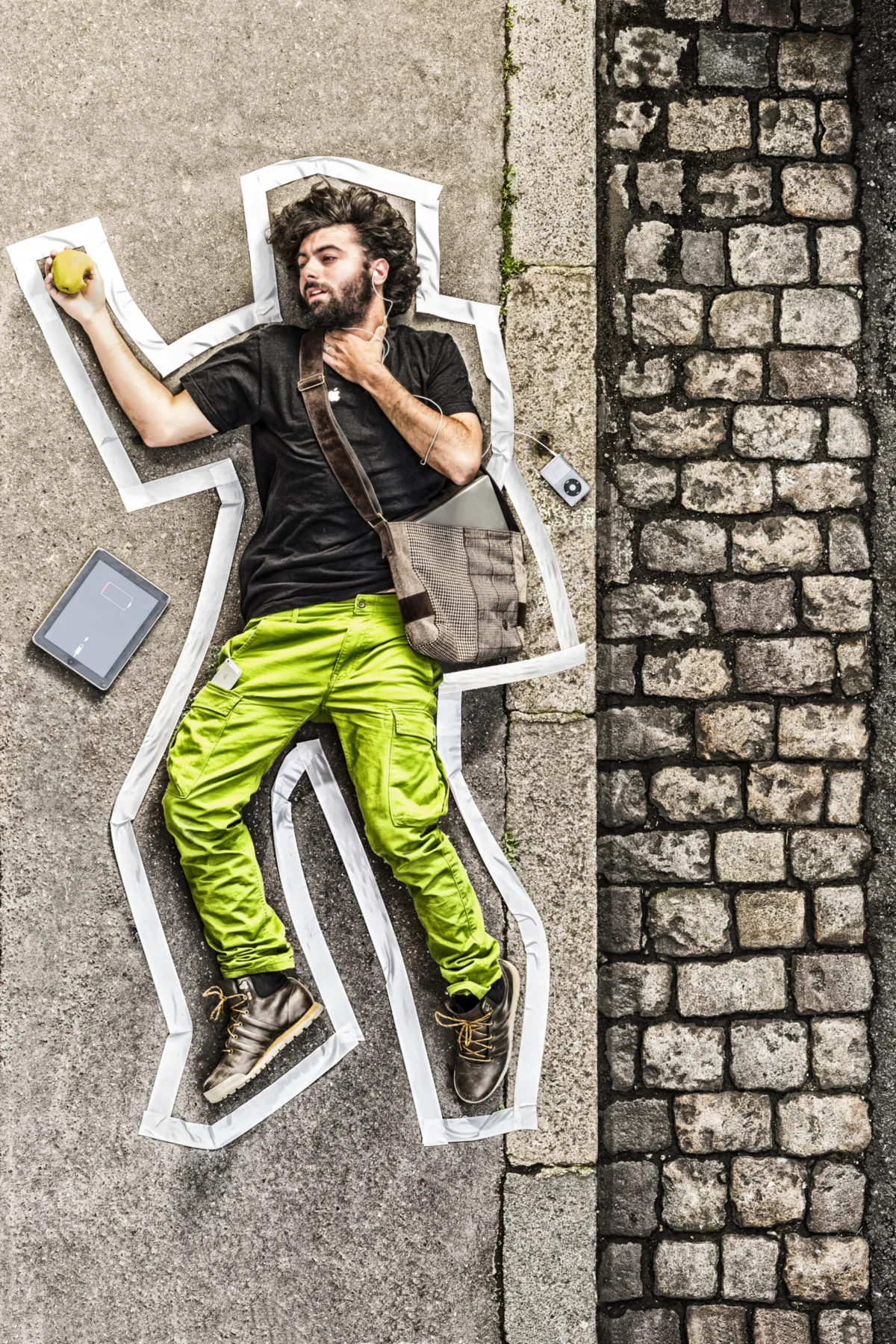 A man in lime trousers lies on cobblestones inside a chalk outline, an apple in hand