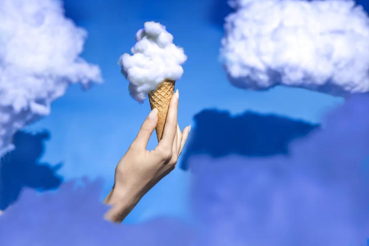 A hand holds an ice-cream cone topped with a cloud against a blue sky