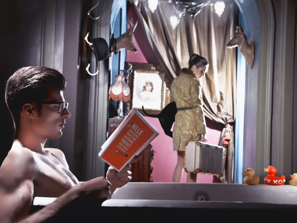 Staged interior: a man reads in a bathtub as a woman in a trench coat passes