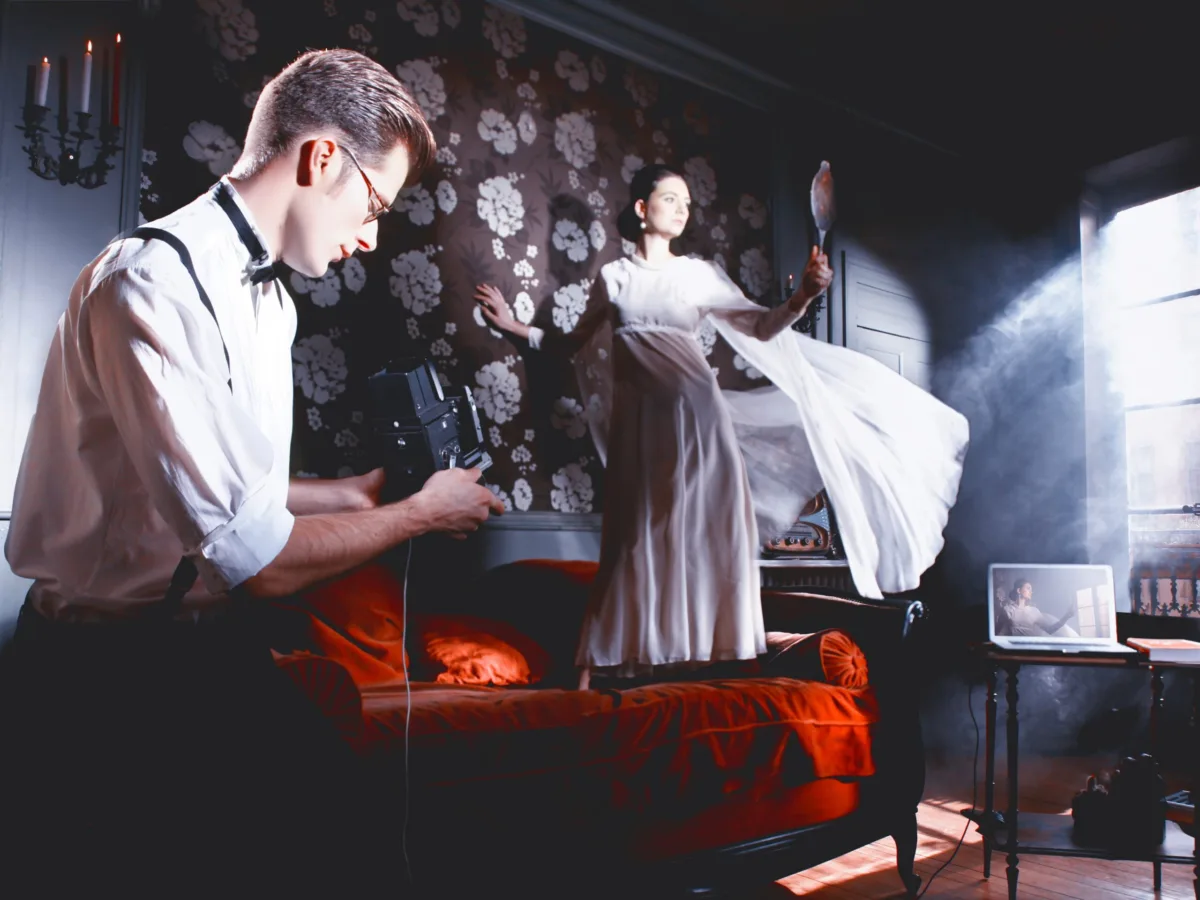 Staged interior: a man reads in a dark room, a woman in a white dress behind him